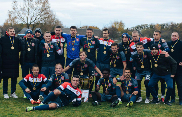 Eine Gruppe von Männern in Medaillen steht auf einem Fußballfeld und hält einen Pokal, umgeben von Bäumen unter einem klaren blauen Himmel.