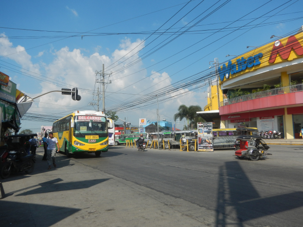 Eine belebte Stadtstraße mit Fahrzeugen wie einem Bus und Motorrädern, Fußgängern auf den Gehwegen, Verkehrsampeln, Strommasten, Gebäuden mit Schildern, Bäumen und einem bewölkten Himmel.