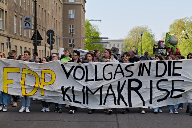 Eine Gruppe von Menschen, die eine Straße entlanggehen und ein Banner mit der Aufschrift "FDP Vollgas in die Klimakrise" tragen, mit Gebäuden, Bäumen und Verkehrszeichen im Hintergrund unter einem klaren blauen Himmel.