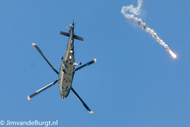 Ein militärisches Helikopter in der Luft, das eine Rakete abfeuert, die Rauch und Feuer ausstößt, mit Text in der unteren linken Ecke.