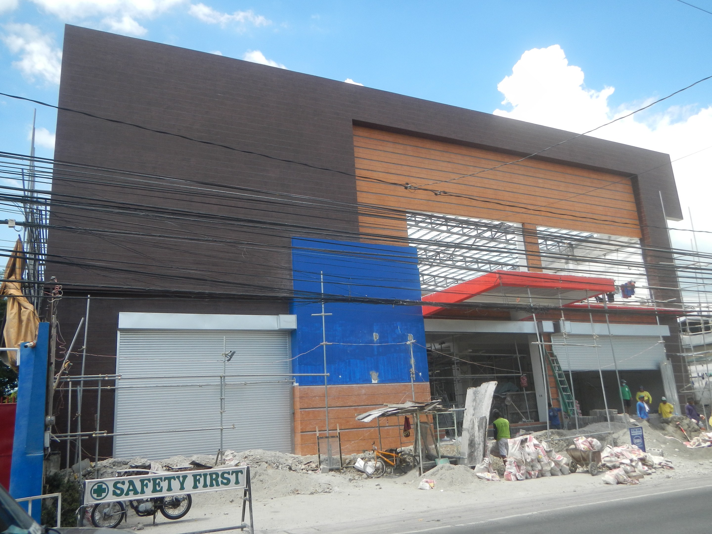 Eine Baustelle mit einem "Sicherheit geht vor"-Schild an einem Gebäude, Fahrzeuge auf der Straße, eine Texttafel, Menschen in der Nähe und Bäume, Pfähle, Drähte und Himmel im Hintergrund.