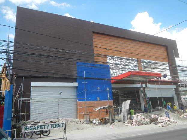 Eine Baustelle mit einem "Sicherheit geht vor"-Schild an einem Gebäude, Fahrzeuge auf der Straße, eine Texttafel, Menschen in der Nähe und Bäume, Pfähle, Drähte und Himmel im Hintergrund.
