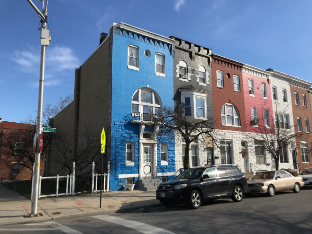 Eine Reihe bunter Häuser an einer Straßenecke in Baltimore, Maryland, mit parkenden Autos, Bäumen, Pfählen, Brettern, einem Zaun und einem bewölkten Himmel im Hintergrund.