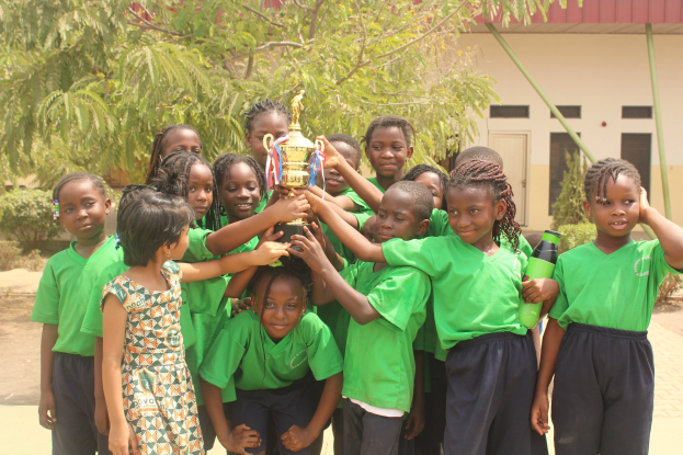 Gruppe von Kindern mit einem Pokal in einem Schulhof mit Bäumen und einem Gebäude im Hintergrund
