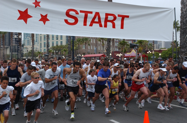 Gruppe von Menschen beim Laufen eines Marathons mit einem Verkehrskegel im Vordergrund und einer Fahne im Hintergrund, umgeben von Bäumen, Laternenmasten, Gebäuden und einem klaren blauen Himmel.