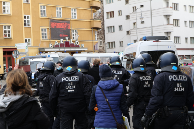 Polizeibeamte in Uniform vor einer Menge bei einer Demonstration in Berlin, mit Fahrzeugen, Gebäuden und einer Person mit Kamera im Bild.