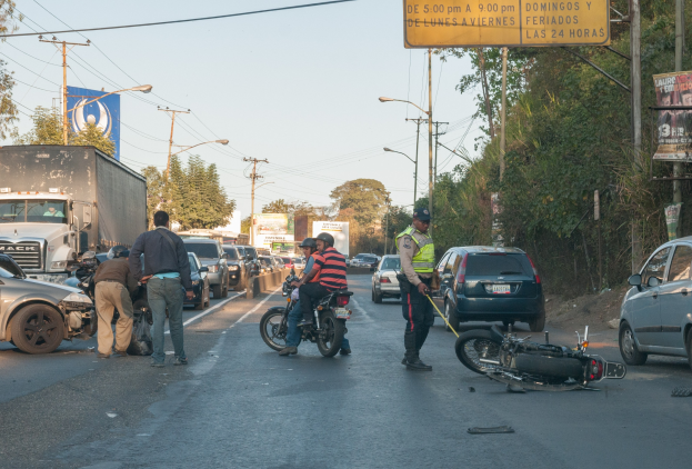 Eine Gruppe von Menschen steht um ein verunglücktes Motorrad am Straßenrand mit mehreren Fahrzeugen, darunter ein Lastwagen, und einer Hintergrund von Bäumen, Masten, Lichtern und Schildern unter dem Himmel.